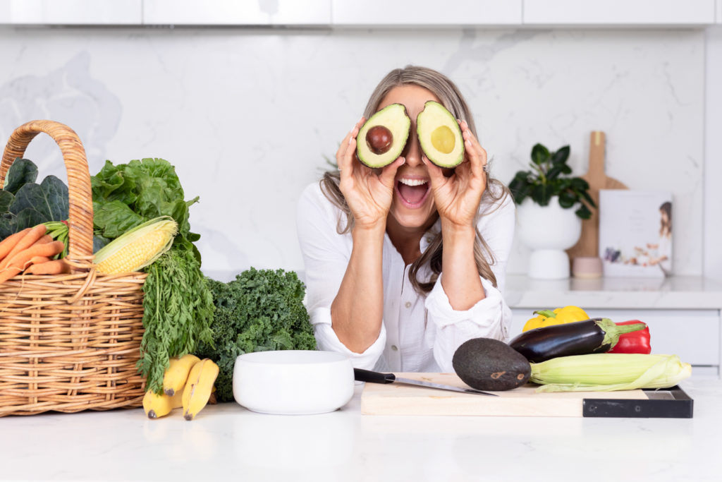 Female Nutritionist leaning at a kitchen bench and holding up two halves of an avocado to cover her eyeso a