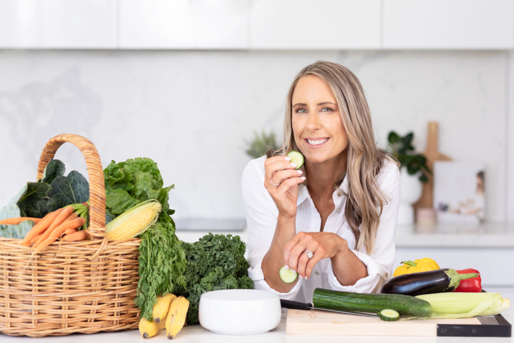 Female Nutritionist leaning at a kitchen bench and eating cucumber with fresh produce around her