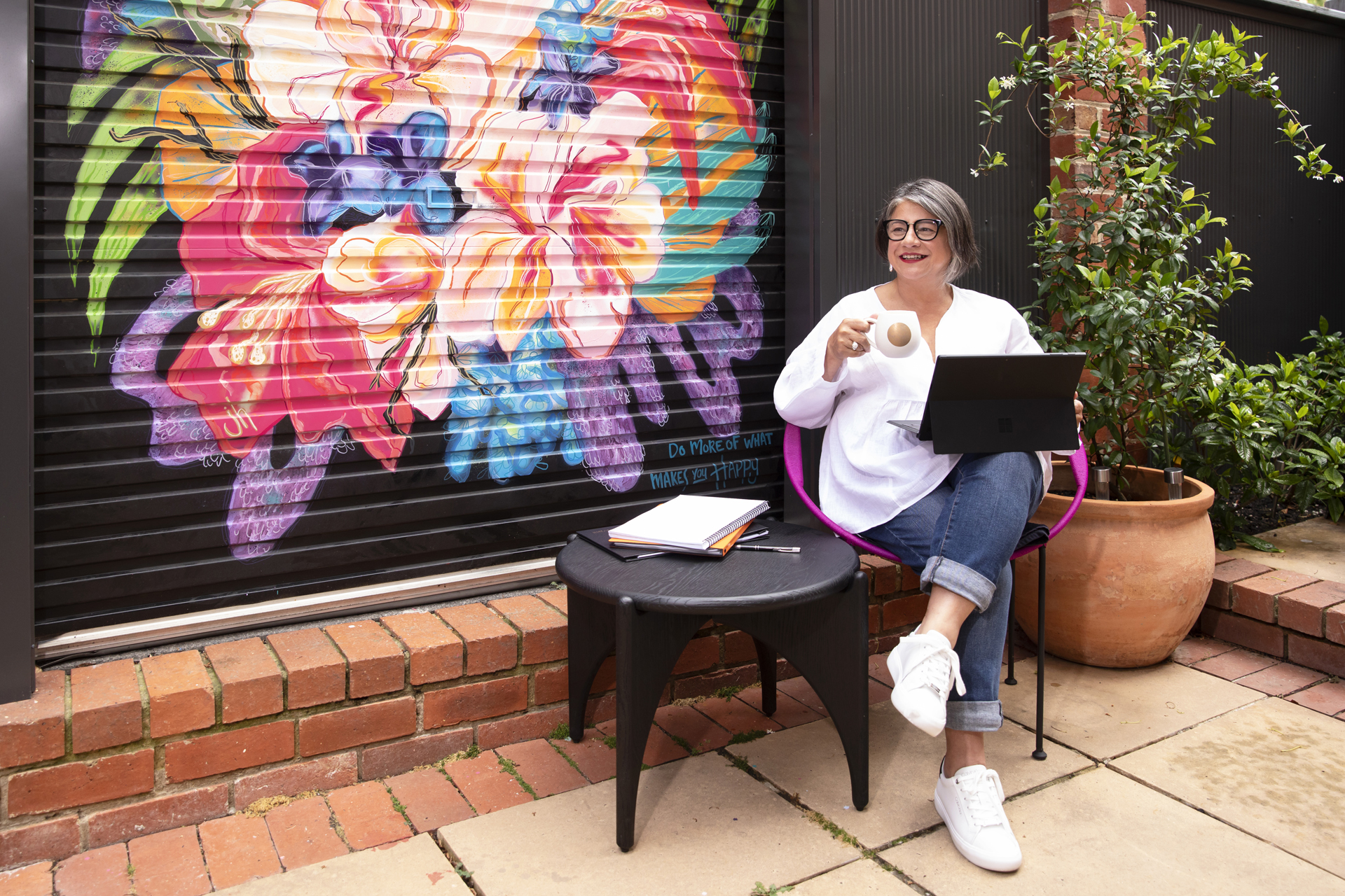 Female business owner holding a cup of tea while working on her laptop in a garden courtyard