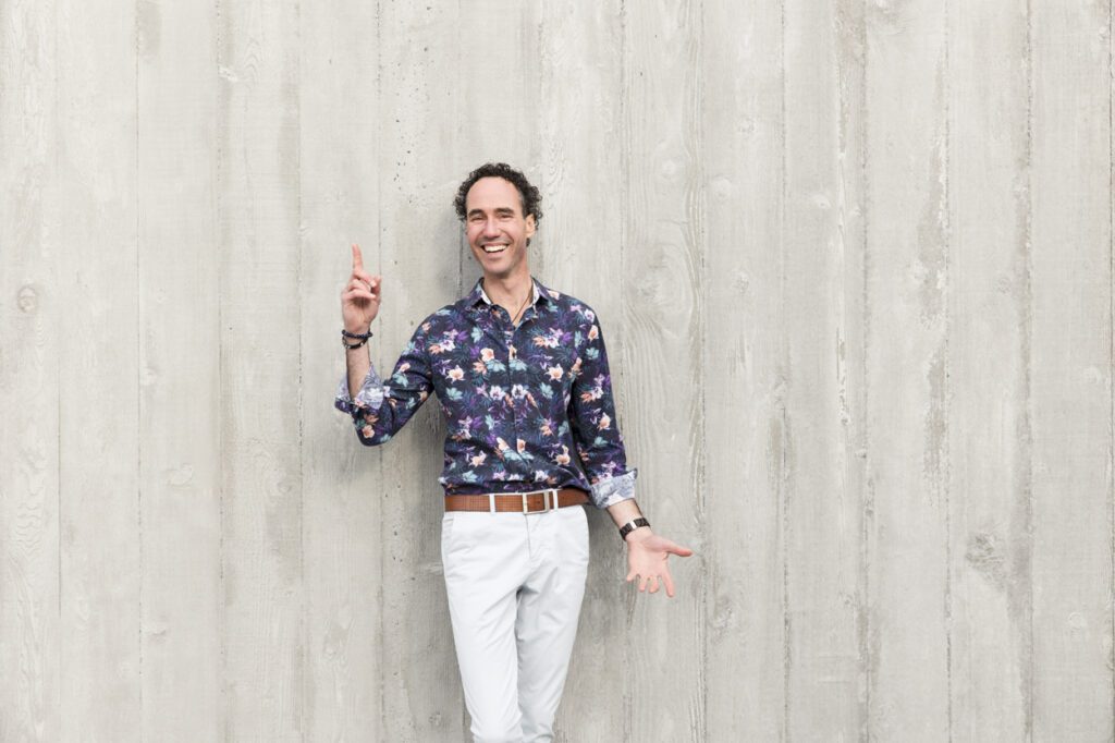 A man standing in front a of a grey wall with a finger pointing upwards