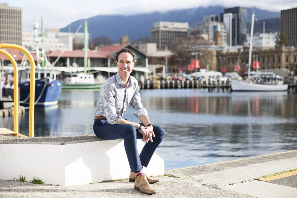 A man sitting down by the dock with boats and city in the background