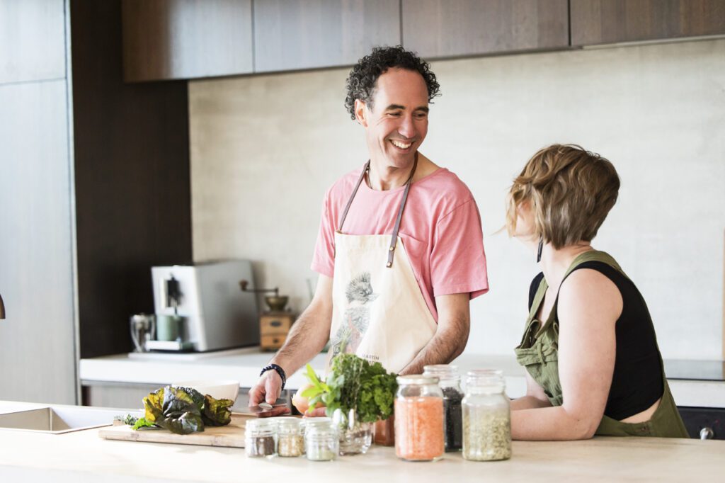 A man talking to a lady while chopping vegetables at the Kitchen bench