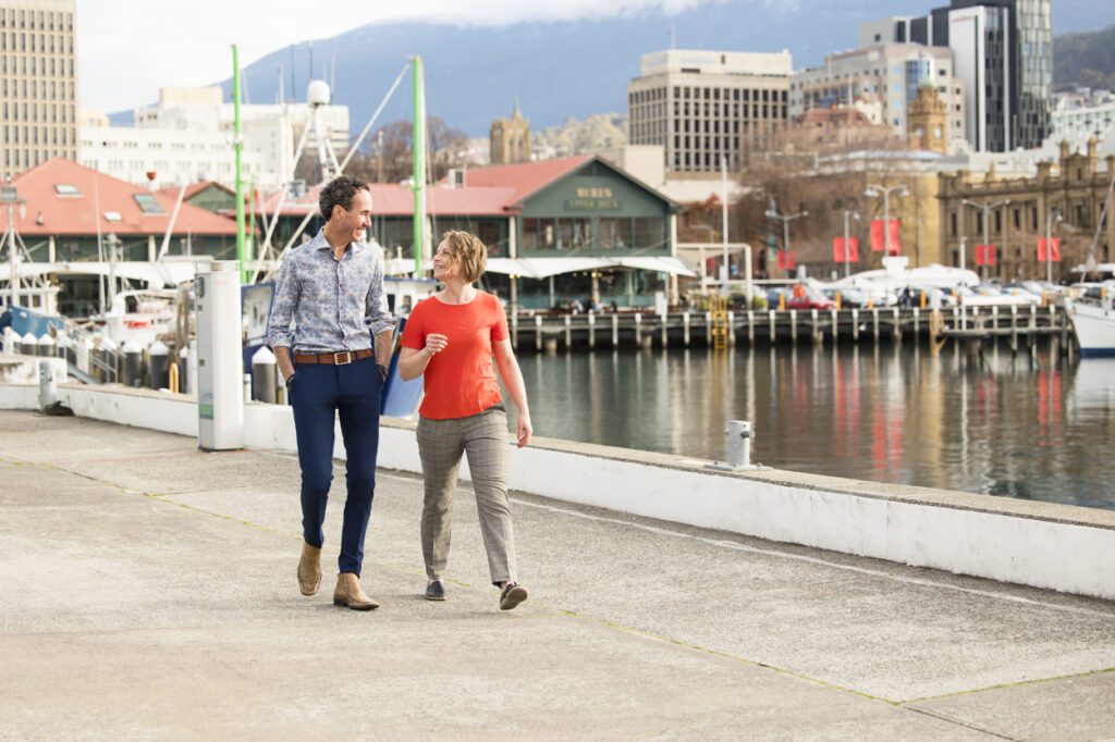 A man and a lady walking together along a dock with boats and city in the background