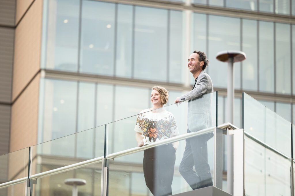 A man and a lady leaning over a balcony with city buildings in the background