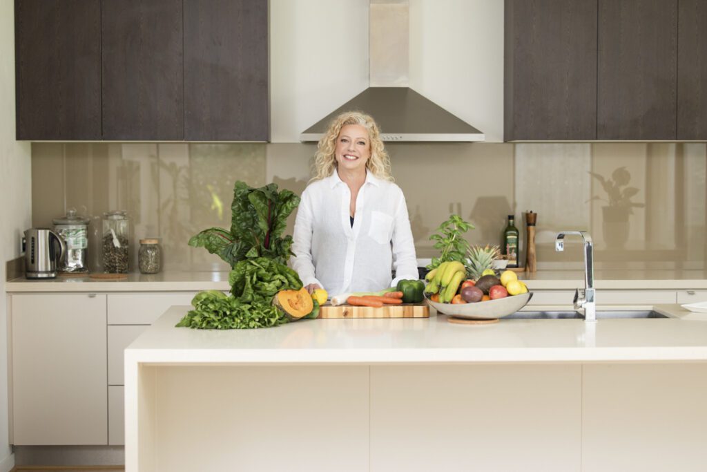 Lady wearing a white shirt standing behind a kitchen bench with fruit and vegetables and a chopping board - personal brand photoshoot
