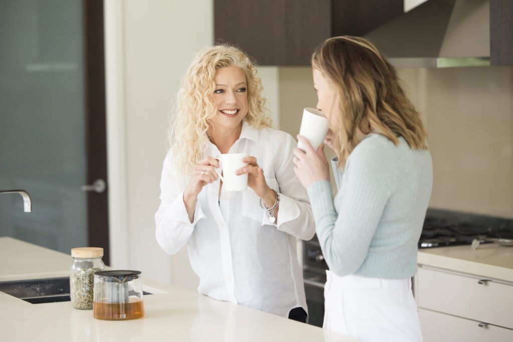 Two ladies, one wearing a white shirt and the other wearing a blue top, drinking tea in a  kitchen - captured on a personal brand photoshoot