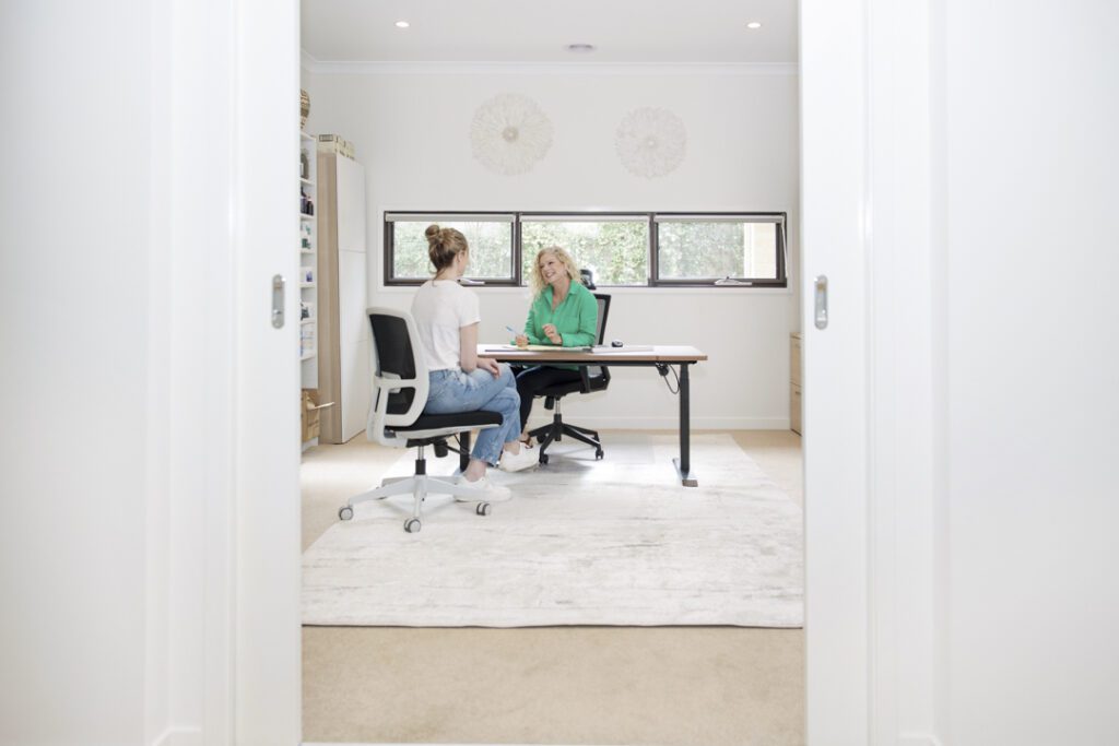 A lady wearing a green shirt sitting at a desk talking to another lady wearing a white-t-shirt who is seated on the opposite side of the desk - captured on a personal brand photoshoot