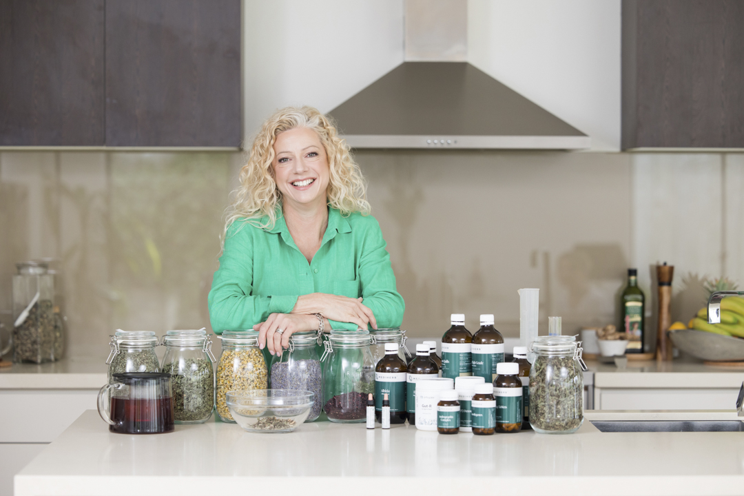 A lady standing at a kitchen bench. There are containers filled with teas and herbs and bottles of oils and health supplements - captured on a personal brand photoshoot