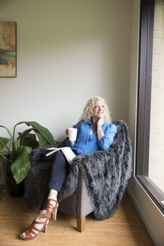 A lady wearing a blue shirt and black pants sitting in a chair holding a cup of tea and looking out the window - captured on a personal brand photoshoot