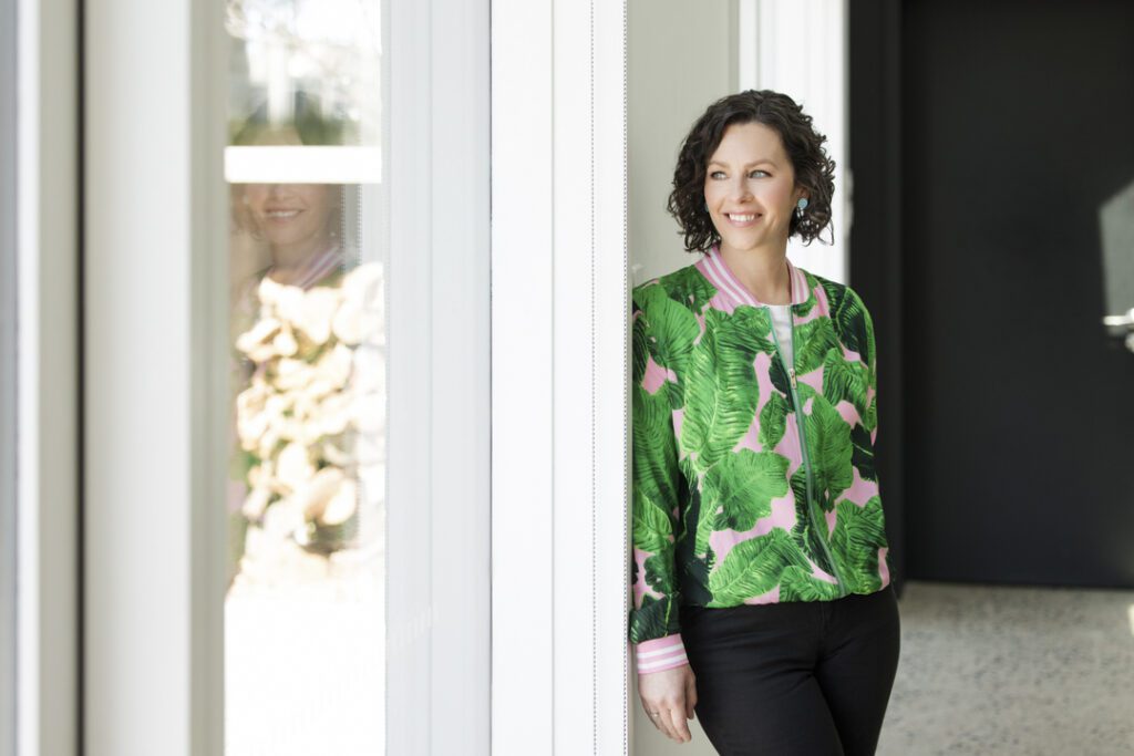 Lady in a cool green and patterned bomber jacket and black jeans leaning against a wall looking out the window