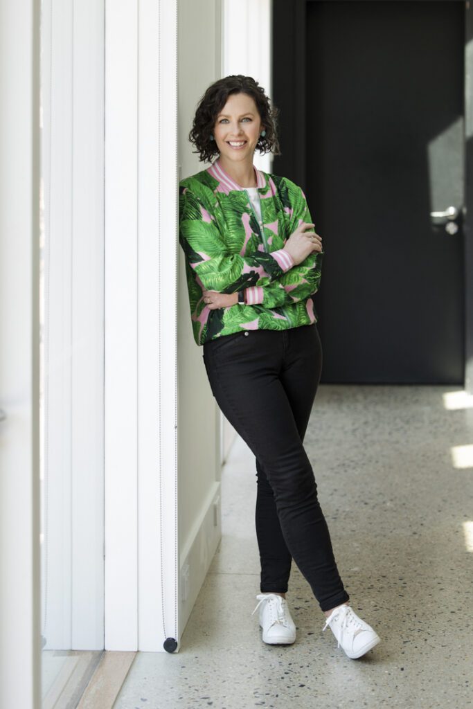 Lady in a green and patterned bomber jacket and black jeans leaning against a wall with one leg crossed over the other