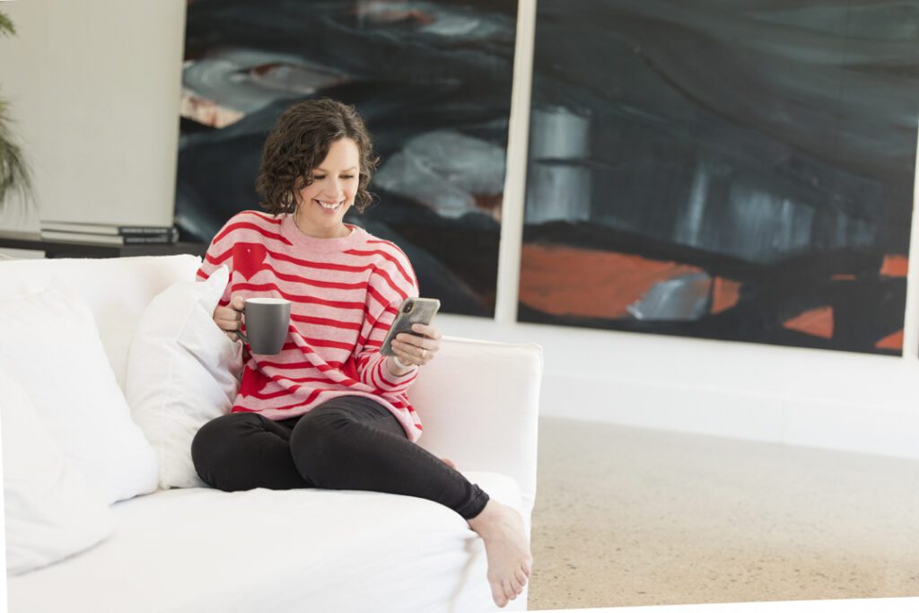 Lady wearing a pink and red stripe jumper and black jeans sitting on a white couch playing on her mobile phone and holding a cup in her other hand