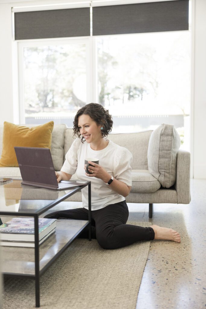 A lady wearing a white top and black jeans sitting on the floor next to a coffee table looking at her laptop screen and holding a cup in her hand