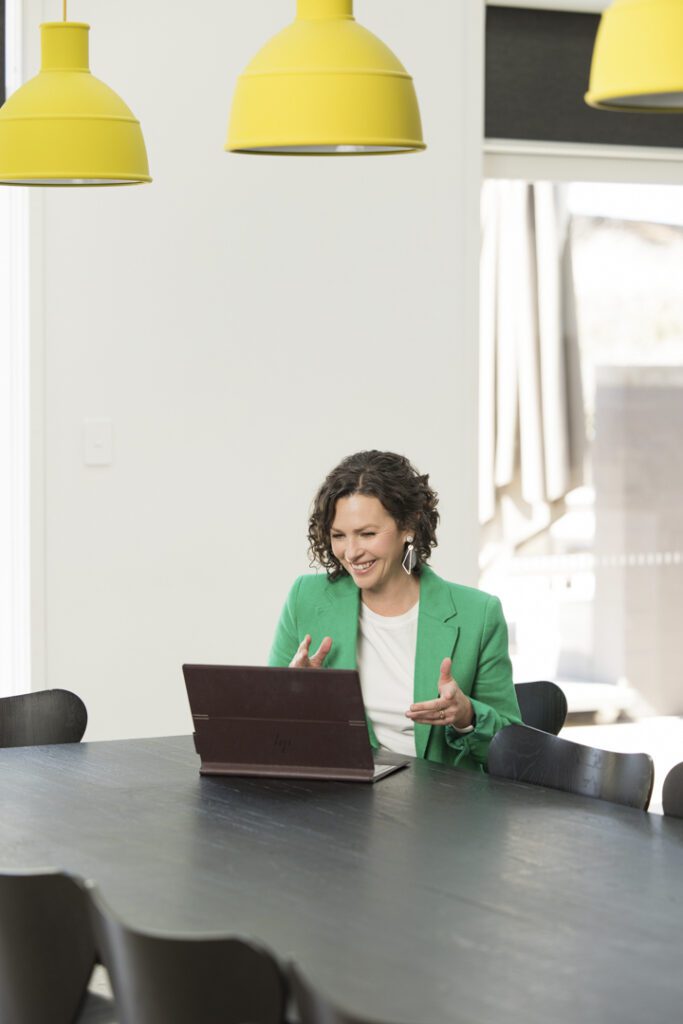 A lady wearing a white top and green jacket sitting at the dinning table looking at her laptop screen