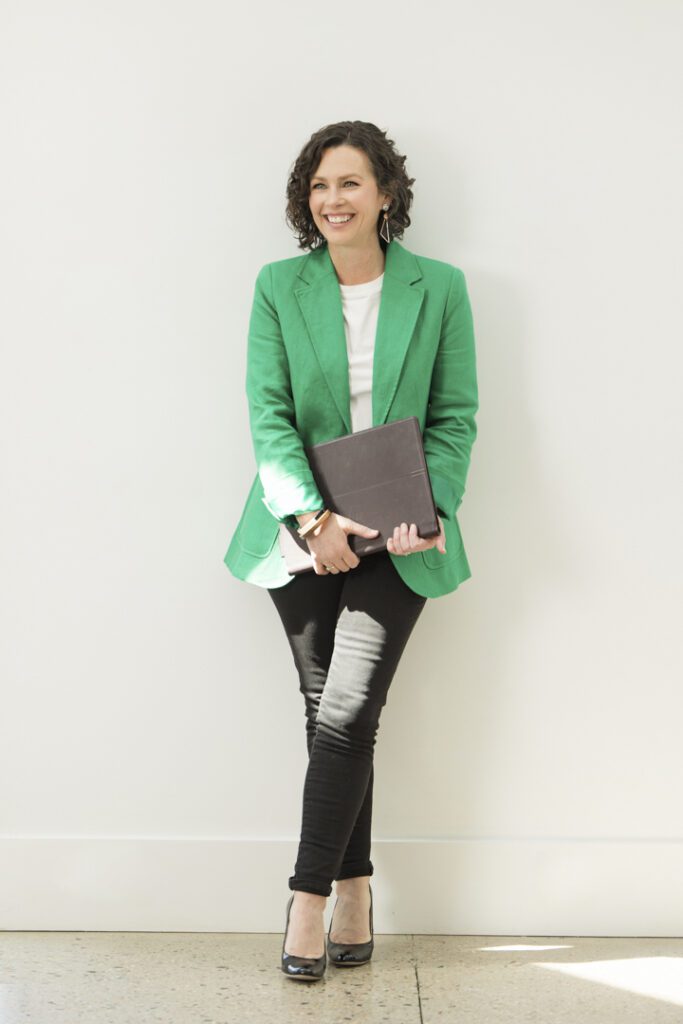 A lady wearing a white top, green jacket and black pants holding a laptop and leaning against a white wall.