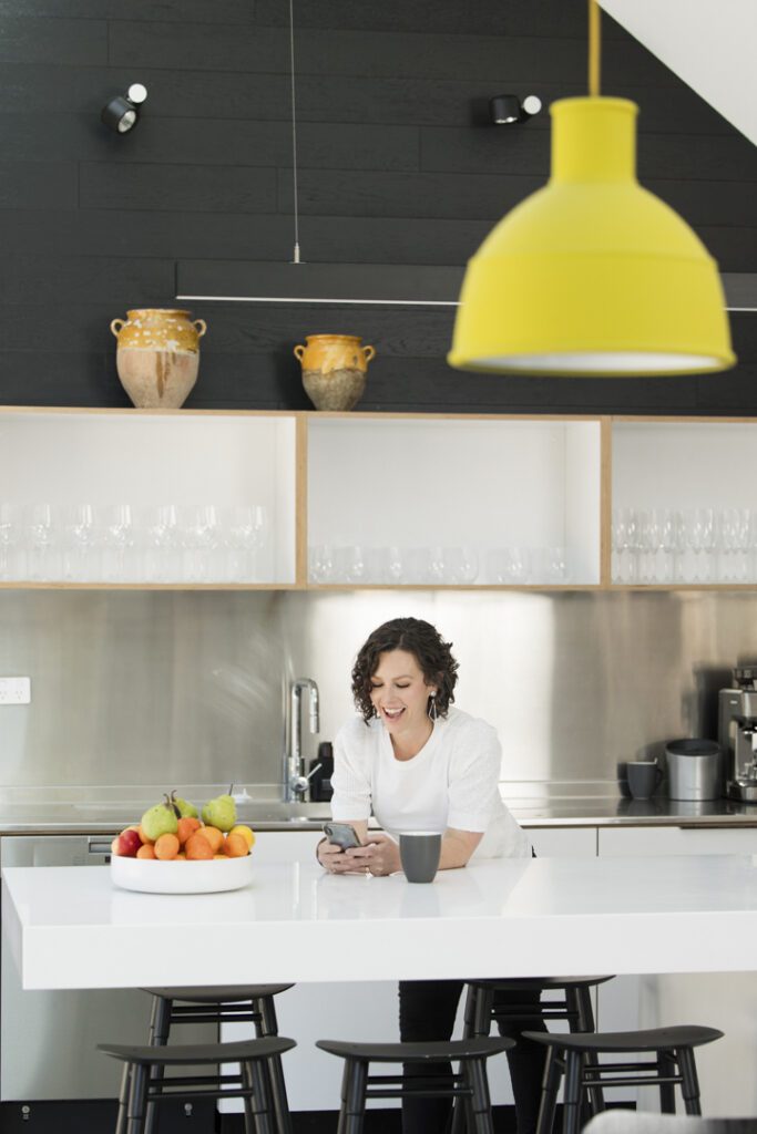 A lady wearing a white top who is leaning over a kitchen bench checking her mobile phone