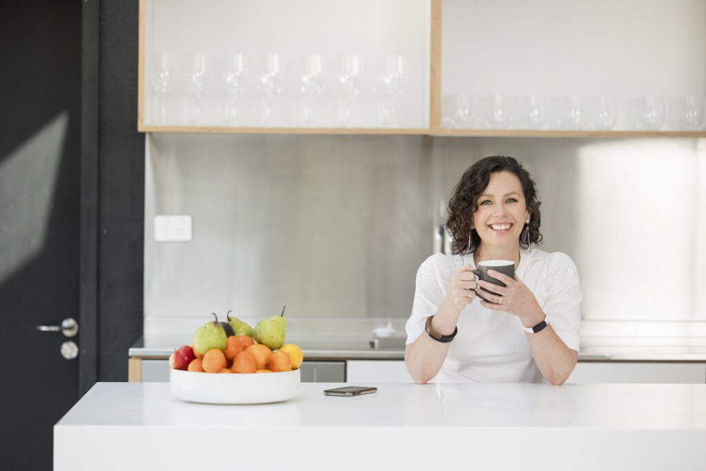 A lady wearing a white top who is sitting at the kitchen bench drinking a cup of tea