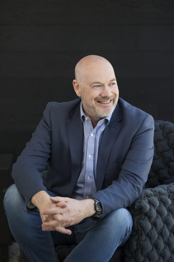 A man wearing a blue shirt and blue jacket and denim jeans sitting in a navy comfy chair in front of a black wall