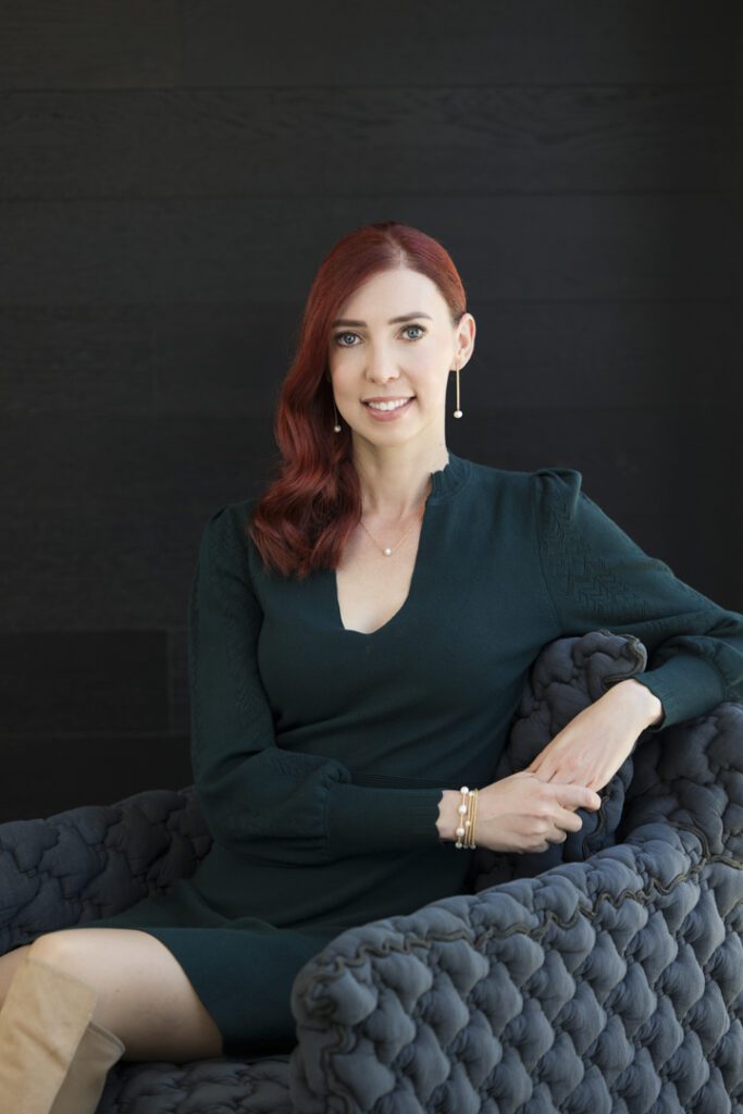 A lady with red hair, wearing a green dress sitting in a navy comfy chair in front of a black wall