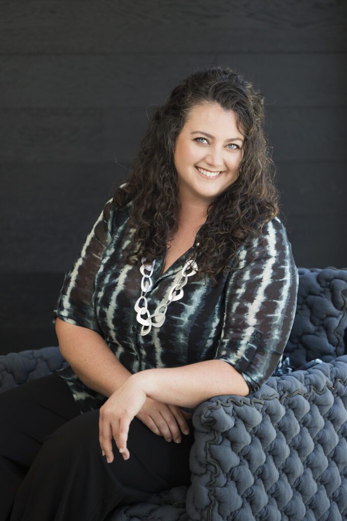 A lady with long curly hair wearing a stripy shirt sitting in a navy blue comfy chair in front of a black wall