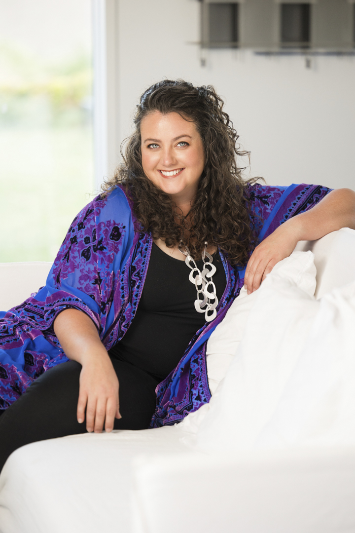 A lady with long curly hair, wearing a black top and black pants with a blue and purple patterned jacket, sitting on a white couch