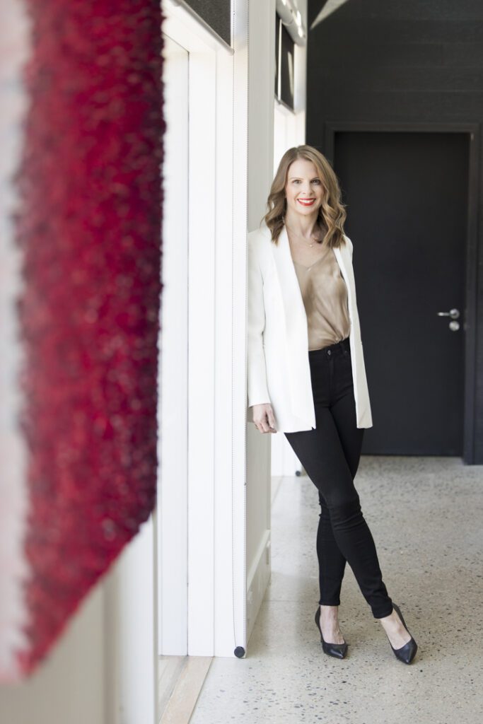 A lady wearing a taupe top and white jacket and black pants leaning against a wall