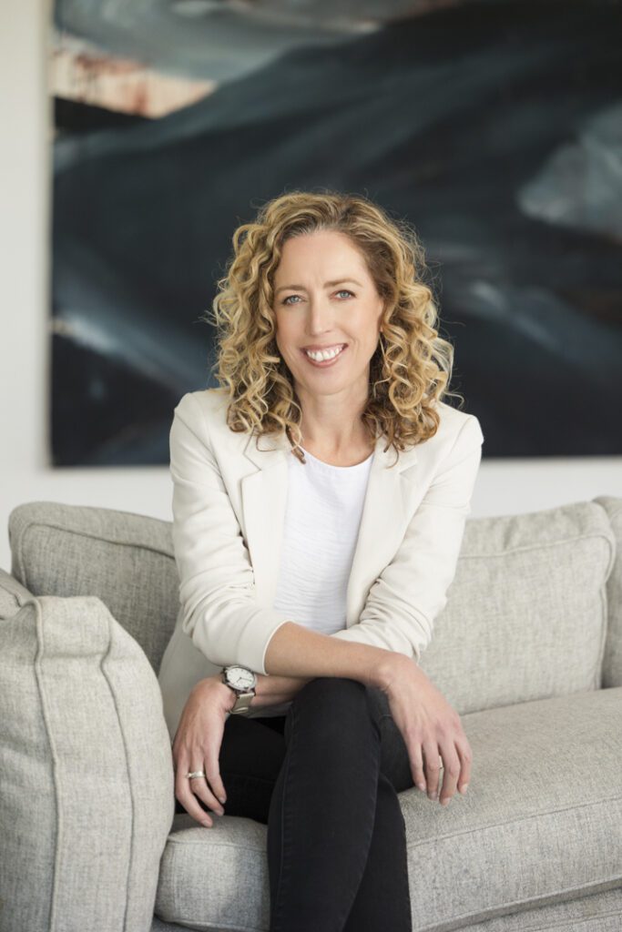 A lady wearing a white top, beige jacket and black pants sitting on a couch