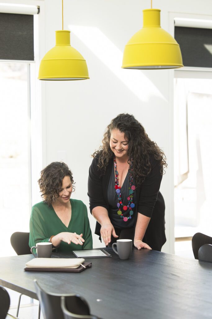 One lady sitting at a table and another standing beside her both looking down at paperwork