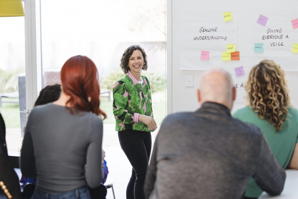 A lady wearing a green patterned jacket and black jeans standing in front a of white board presenting to her work colleagues