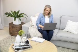 A portrait of a lady wearing a blue shirt and jeans sitting. on her lounge as she works on her laptop