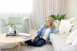 A personal brand photoshoot that captures a portrait of a lady wearing a blue shirt and jeans sitting on the floor of her lounge room  and leaning against the side of her lounge.