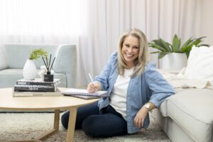 A personal brand photoshoot that captures a portrait of a lady wearing a blue shirt and jeans sitting on the floor of her lounge room  writing in her notebook which is on the coffee table