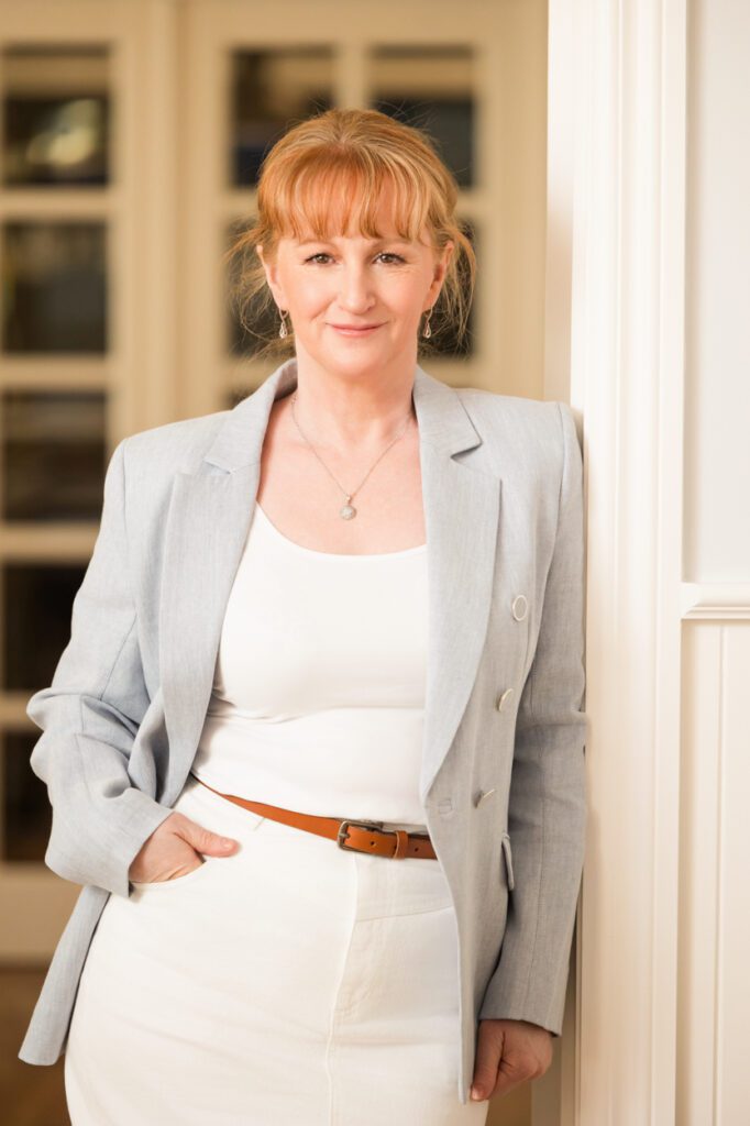 A lady wearing a white top and a light blue jacket leaning against a wall