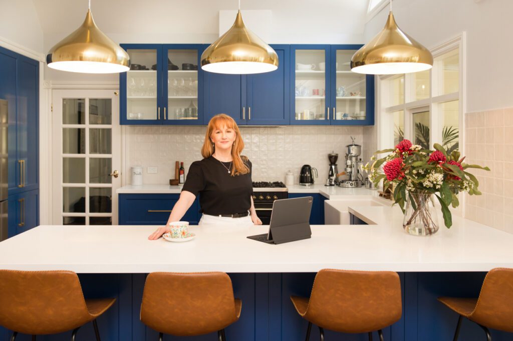 A lady wearing a black t-shirt leaning over a kitchen bench with blue cupboard doors as she is drinking a cup of tea and looking at her lap top