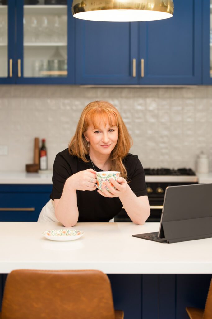 A lady wearing a black t-shirt leaning over a kitchen bench with blue cupboard doors as she is drinking a cup of tea and looking at her lap top