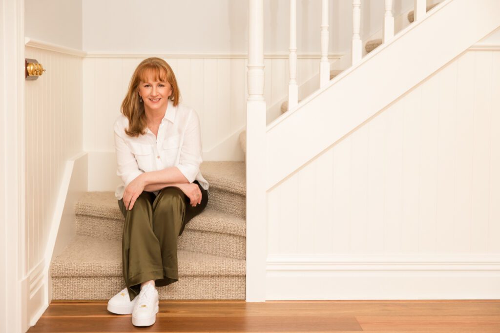 A lady wearing a white linen shirt sitting on the bottom steps of stairs in a house