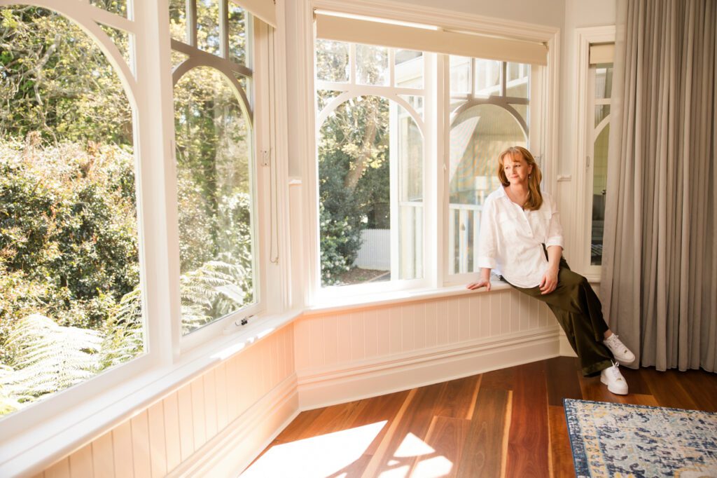 A lady wearing a white linen shirt sitting on a window sill looking out into a garden