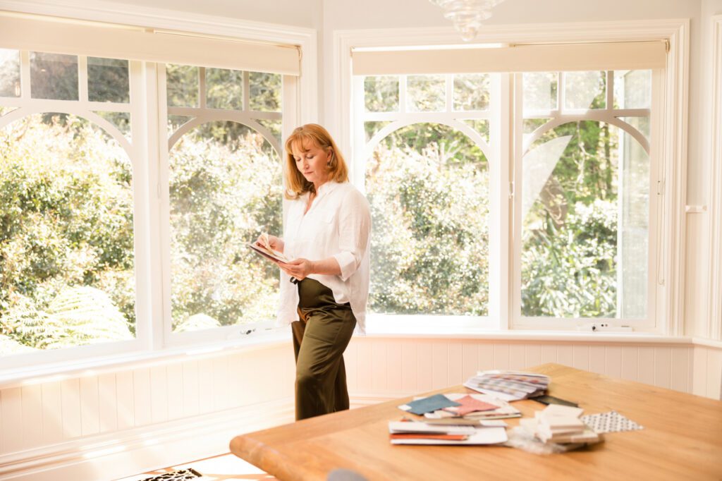 An Interior designer standing in front of windows in a heritage house looking at a colour wheel she is holding. There are other samples and swatches on the table in the foreground