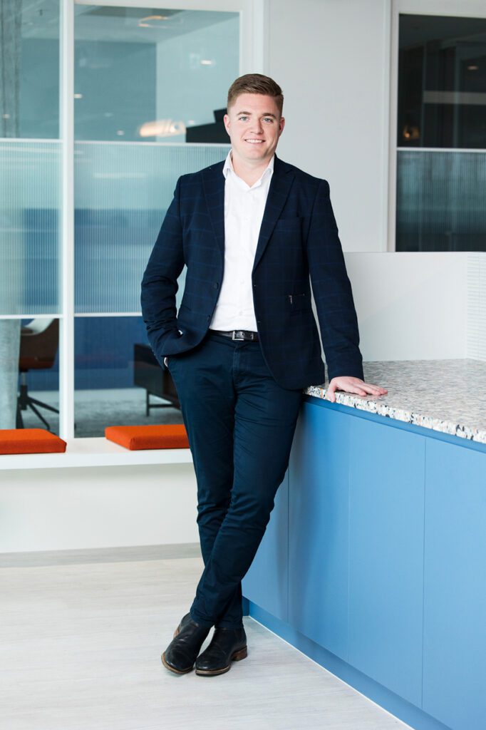 Full length photos of a man in a navy suit, leaning against a bench with one hand in his pocket