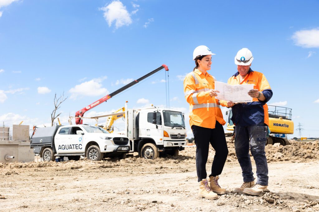 A female and a male wearing high vis clothing and hard hats looking over work plans with a worksite and work vehicle behind them