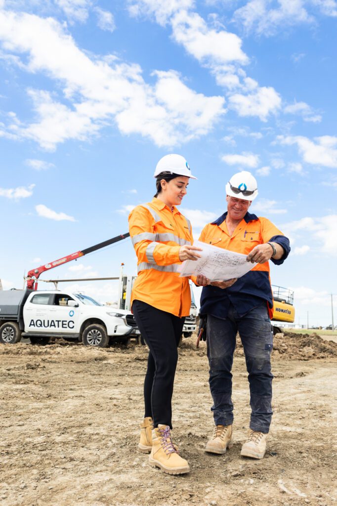 A female and a male wearing high vis clothing and hard hats looking over work plans with a worksite and work vehicle behind them