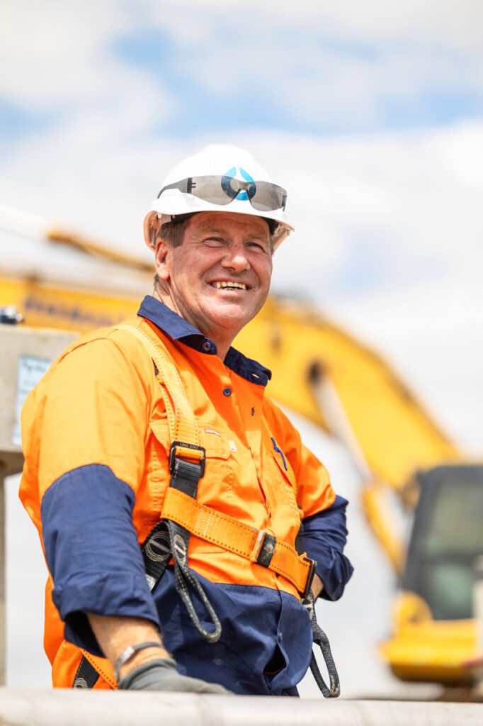 A man wearing high vis clothing and a hard hat with a worksite behind him