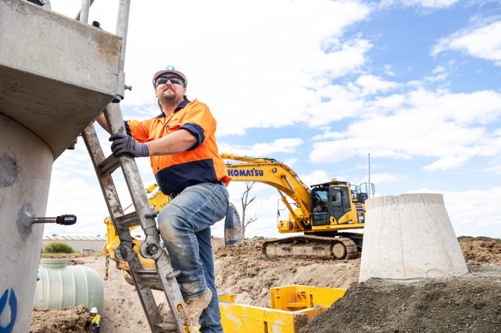 A man wearing high vis clothing and a hard hat climbing a ladder with a worksite and bulldozer behind him