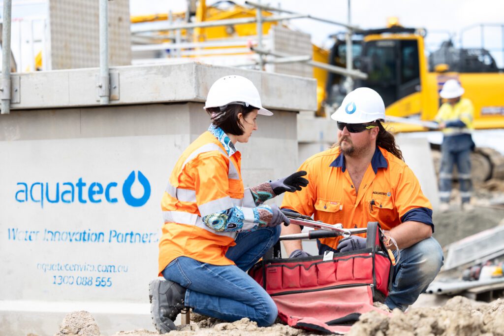 A female and a male wearing high vis clothing and hard hats getting tools out of a tool box with a worksite and and machinery behind them