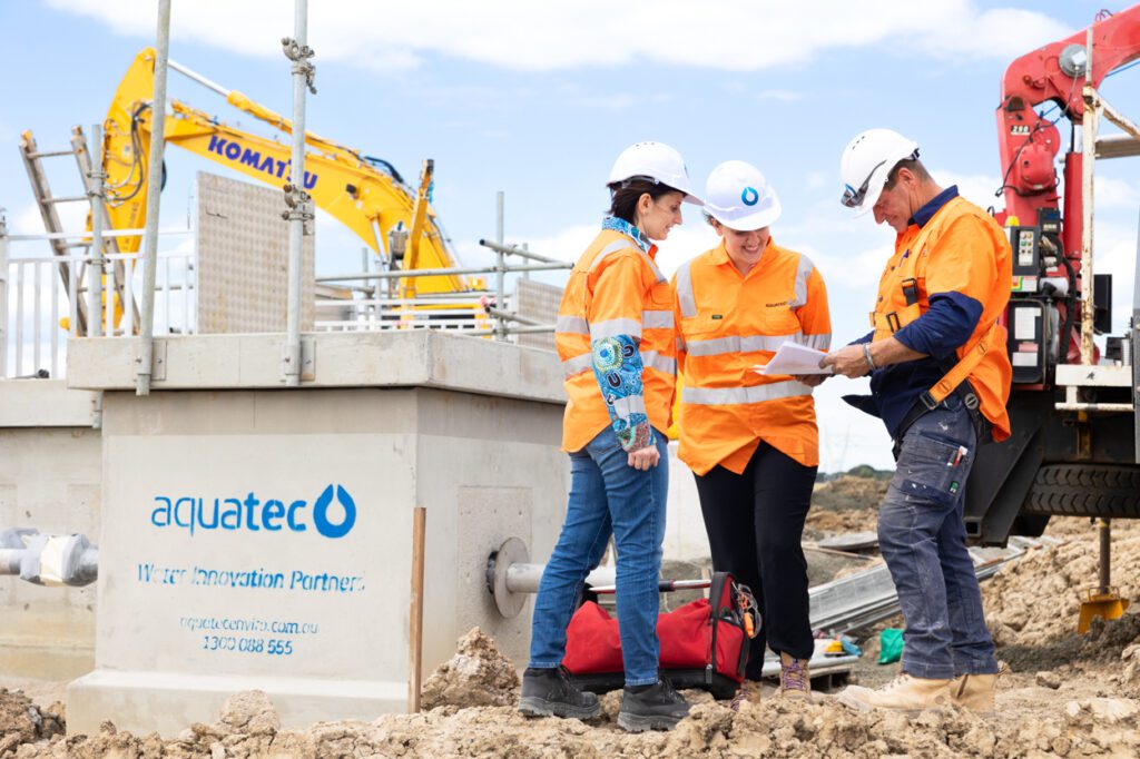 Two females and a male wearing high vis clothing and hard hats looking over work plans with a worksite and and machinery behind them