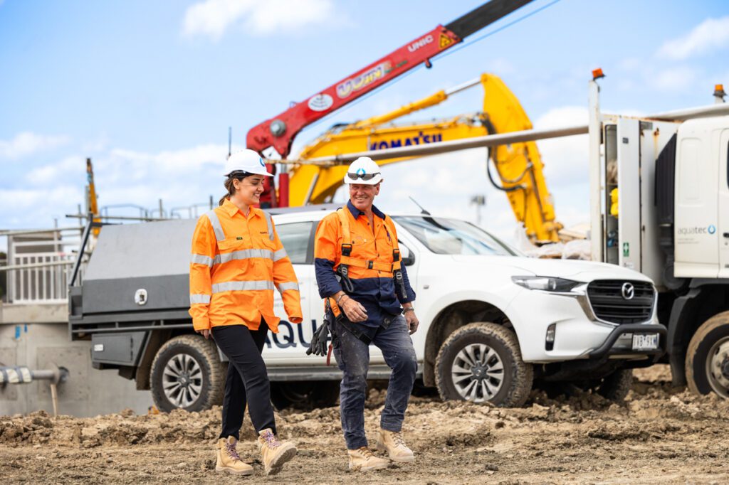 A female and a male wearing high vis clothing and hard hats walking and talking to each other with a worksite, work vehicles and machinery behind them