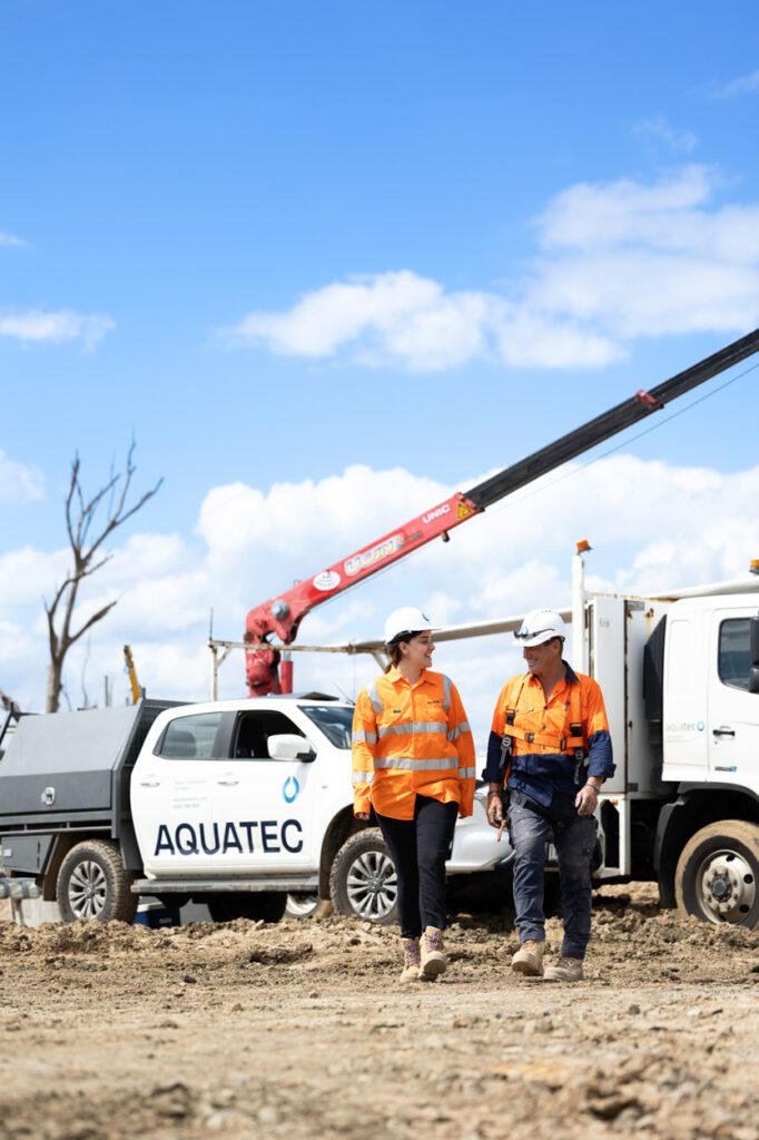 A female and a male wearing high vis clothing and hard hats walking and talking to each other with a worksite and work vehicle behind them