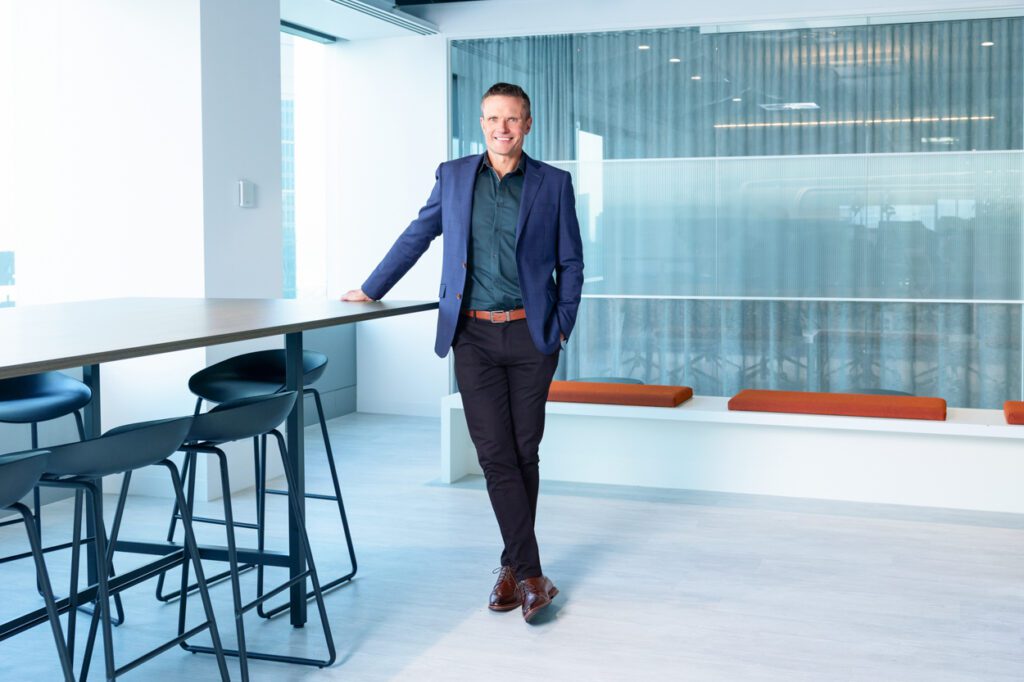 Full length portrait of a man in an office wearing black pants, a green shirt and a blue blazer