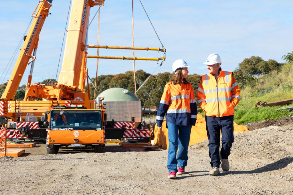 A male and a female wearing high vis clothing and hard hats standing walking and talking with a worksite in the background