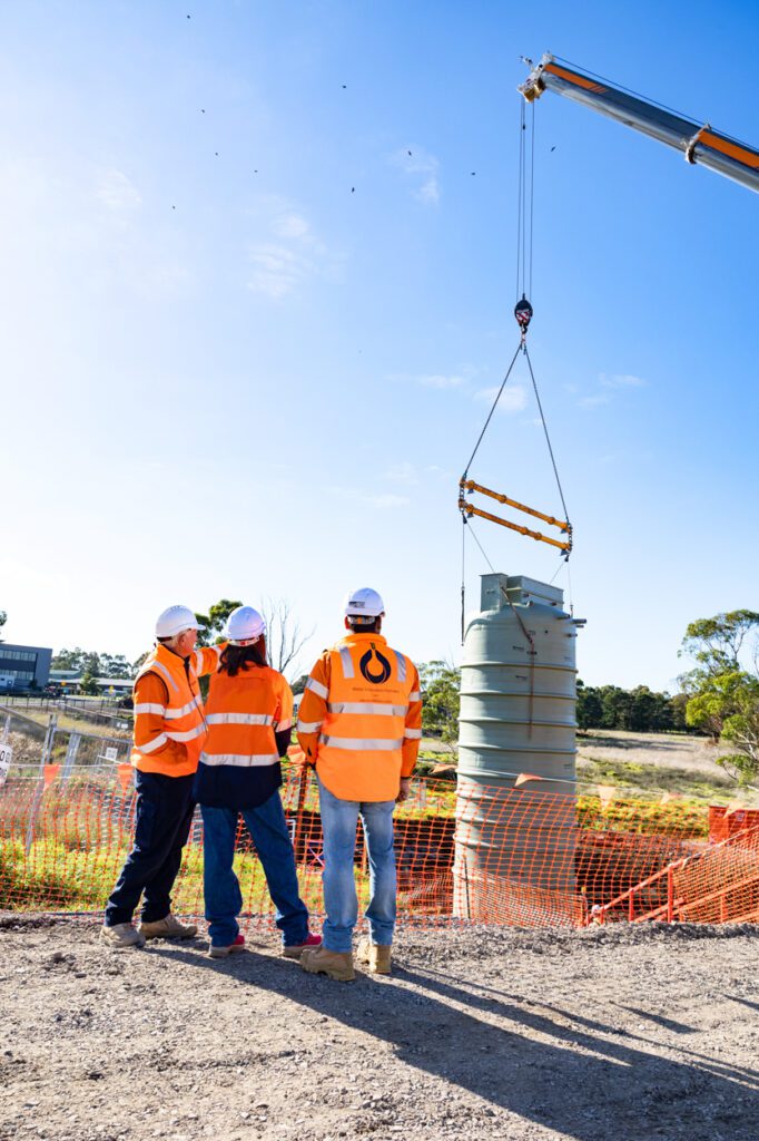 Two males and one female wearing high vis clothing and hard hats standing gathered at a worksite as a water storage tank is being lowered into the ground by a crane